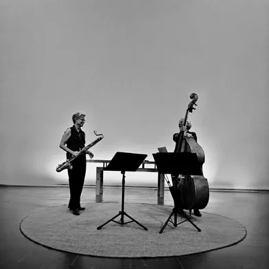 Schwarz-Weiß-Foto zweier hellhäutiger Frauen bei einem musikalischen Auftritt. Links steht eine Saxofonistin, rechts eine Kontrabassistin. Vor ihnen stehen zwei Notenpulte. Der schlichte, helle Raum und der runde Teppich bilden die Kulisse.