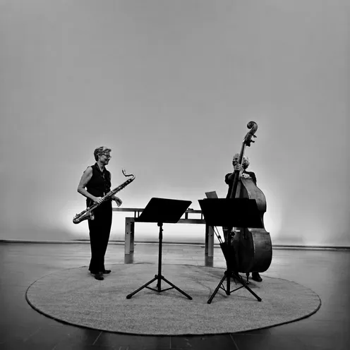 Schwarz-Weiß-Foto zweier hellhäutiger Frauen bei einem musikalischen Auftritt. Links steht eine Saxofonistin, rechts eine Kontrabassistin. Vor ihnen stehen zwei Notenpulte. Der schlichte, helle Raum und der runde Teppich bilden die Kulisse.