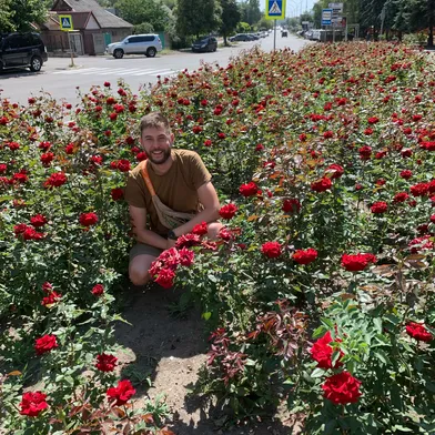 Ein hellhäutiger Mann mit Bart kniet lächelnd inmitten eines üppigen Beets voller roter Rosen. Er trägt ein braunes T-Shirt und Shorts. Im Hintergrund verlaufen eine sonnige Straße mit Autos, Häusern, Bäumen und Zebrastreifen.