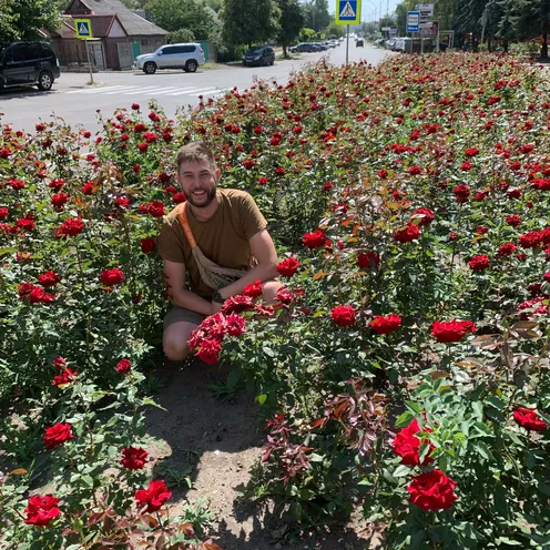 Ein hellhäutiger Mann mit Bart kniet lächelnd inmitten eines üppigen Beets voller roter Rosen. Er trägt ein braunes T-Shirt und Shorts. Im Hintergrund verlaufen eine sonnige Straße mit Autos, Häusern, Bäumen und Zebrastreifen.