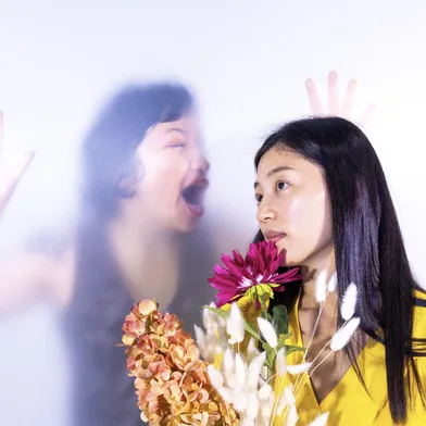 Zwei südkoreanische Frauen, eine im Vordergrund mit gelbem Oberteil und Blumen, die andere im Hintergrund hinter einer milchigen Scheibe mit geöffnetem Mund und erhobener Hand