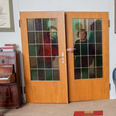 Zwei hellhäutige Männer stehen hinter einer doppelflügeligen Holztür mit bunten Glasscheiben. Einer trägt eine rote Jacke, der andere dunkle Kleidung. Im gemütlichen Wohnzimmer stehen ein Klavier, ein Hocker und ein blaues Sofa.