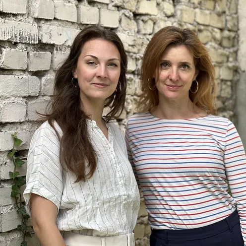 Zwei hellhäutige Frauen stehen vor einer alten, teilweise verputzten Backsteinwand. Eine hat lange dunkle Haare und eine helle Bluse, die andere rötliche Haare und ein weißes, rot-blau gestreiftes Oberteil. Beide blicken in die Kamera.