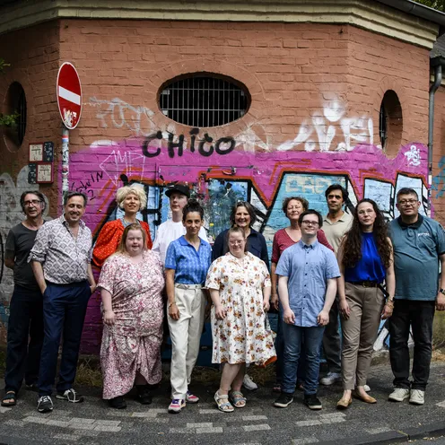Eine Gruppe aus Frauen und Männern mit überwiegend heller, teils mittlerer Hautfarbe posiert in bunter Sommerkleidung vor einer Backsteinwand mit farbenfrohen Graffiti. Die Personen stehen in zwei Reihen auf einem Gehweg.