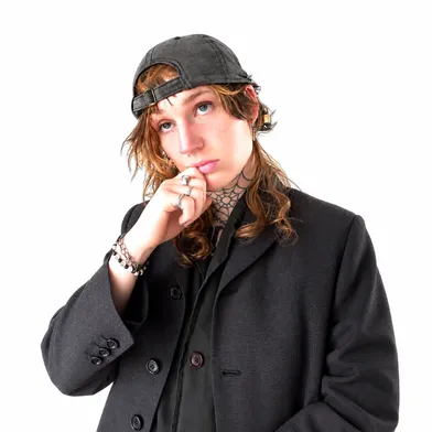 Porträt einer Person mit rückwärts getragener schwarzer Kappe und langen braunen Haaren. Sie trägt einen schwarzen Blazer, Schmuck und Piercings sowie ein Spinnennetz-Tattoo am Hals. Eine Hand liegt nachdenklich am Kinn.