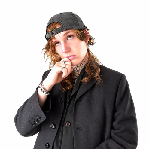 Porträt einer Person mit rückwärts getragener schwarzer Kappe und langen braunen Haaren. Sie trägt einen schwarzen Blazer, Schmuck und Piercings sowie ein Spinnennetz-Tattoo am Hals. Eine Hand liegt nachdenklich am Kinn.